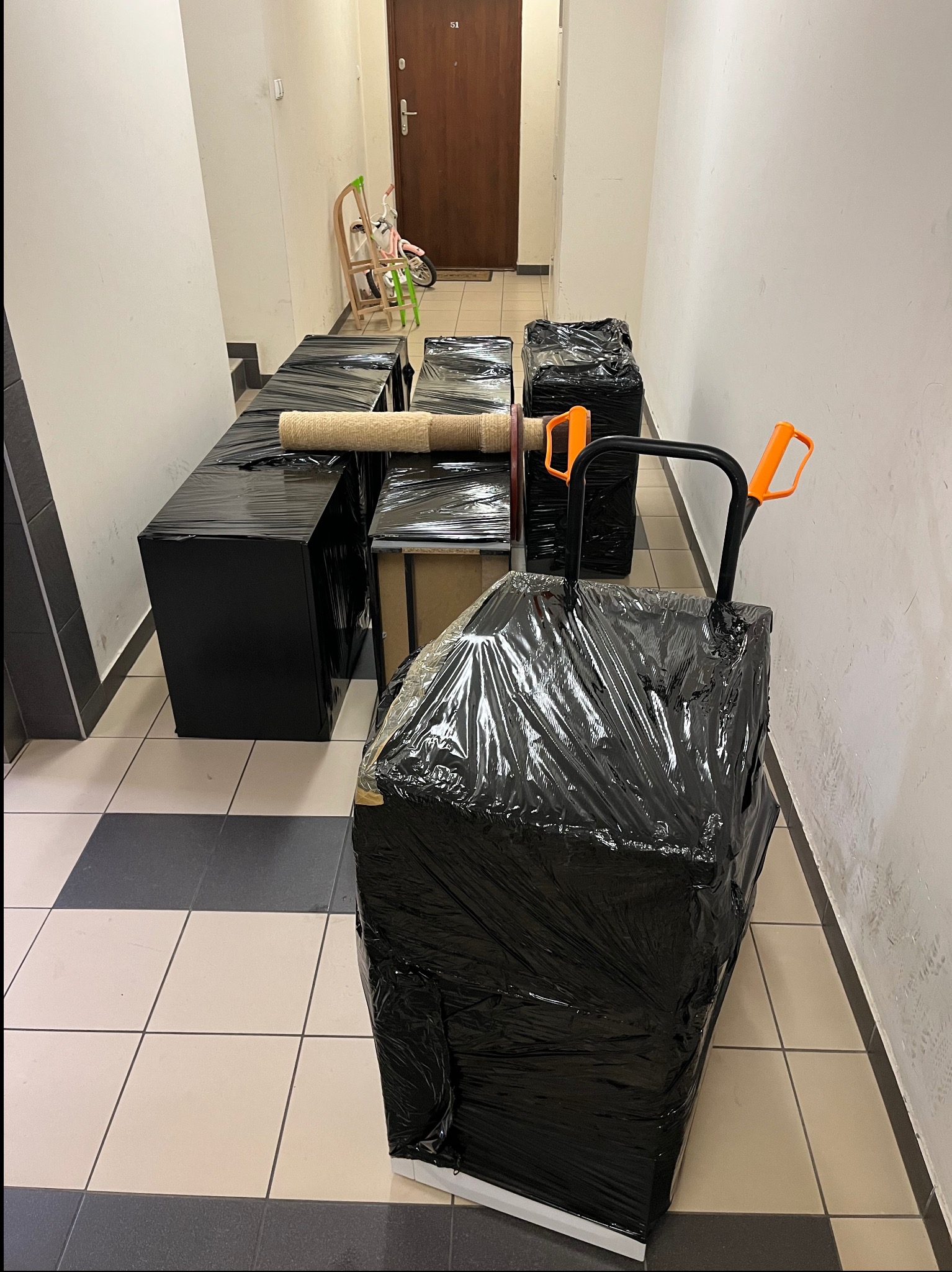 Meble i inne przedmioty zabezpieczone folią i przygotowane do transportu, ustawione w korytarzu bloku mieszkalnego, widoczny wózek transportowy z pomarańczowymi uchwytami.