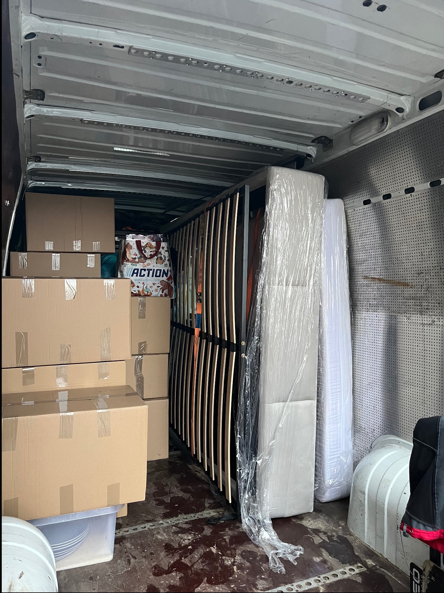 Wnętrze furgonetki wypełnionej kartonami, stelażem łóżka zabezpieczonym pasami i materacami w folii, torba z logo Action widoczna między kartonami.