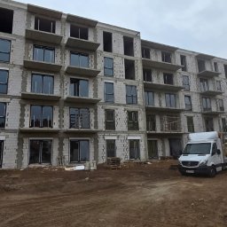 QFENSTER Jan Kowalski - Montaż stolarki okiennej w bloku mieszkalnym w Mostkowie, widoczne okna z czarnymi ramami, budynek w stanie surowym, na placu budowy stoi samochód dostawczy i dźwig.