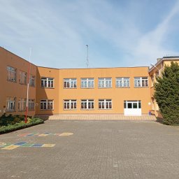 QFENSTER Jan Kowalski - Żółty budynek Szkoły Podstawowej w Przelewicach z nowymi oknami, widok z zewnątrz na dziedziniec z namalowanymi grami podwórkowymi.