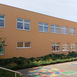 QFENSTER Jan Kowalski - Fasada budynku w ciepłym, pomarańczowym odcieniu z licznymi białymi oknami, niektóre ozdobione papierowymi sercami, otoczona zielenią krzewów i drzew. Na pierwszym planie chodnik z namalowanymi...