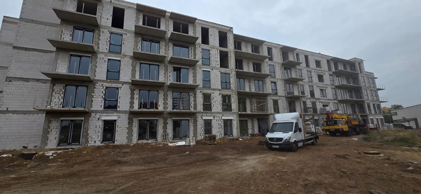 Montaż stolarki okiennej w bloku mieszkalnym w Mostkowie, widoczne okna z czarnymi ramami, budynek w stanie surowym, na placu budowy stoi samochód dostawczy i dźwig.