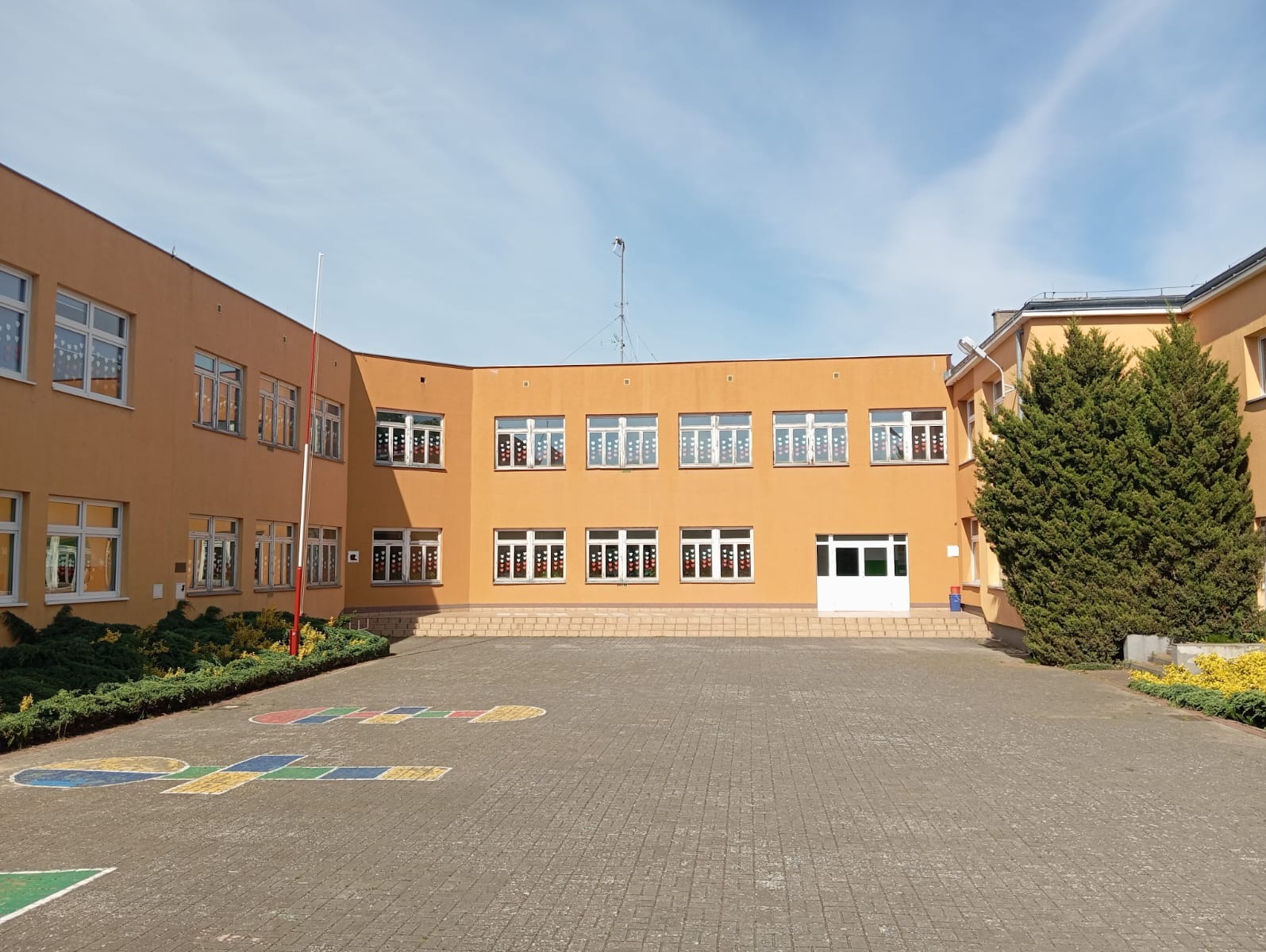 Żółty budynek Szkoły Podstawowej w Przelewicach z nowymi oknami, widok z zewnątrz na dziedziniec z namalowanymi grami podwórkowymi.