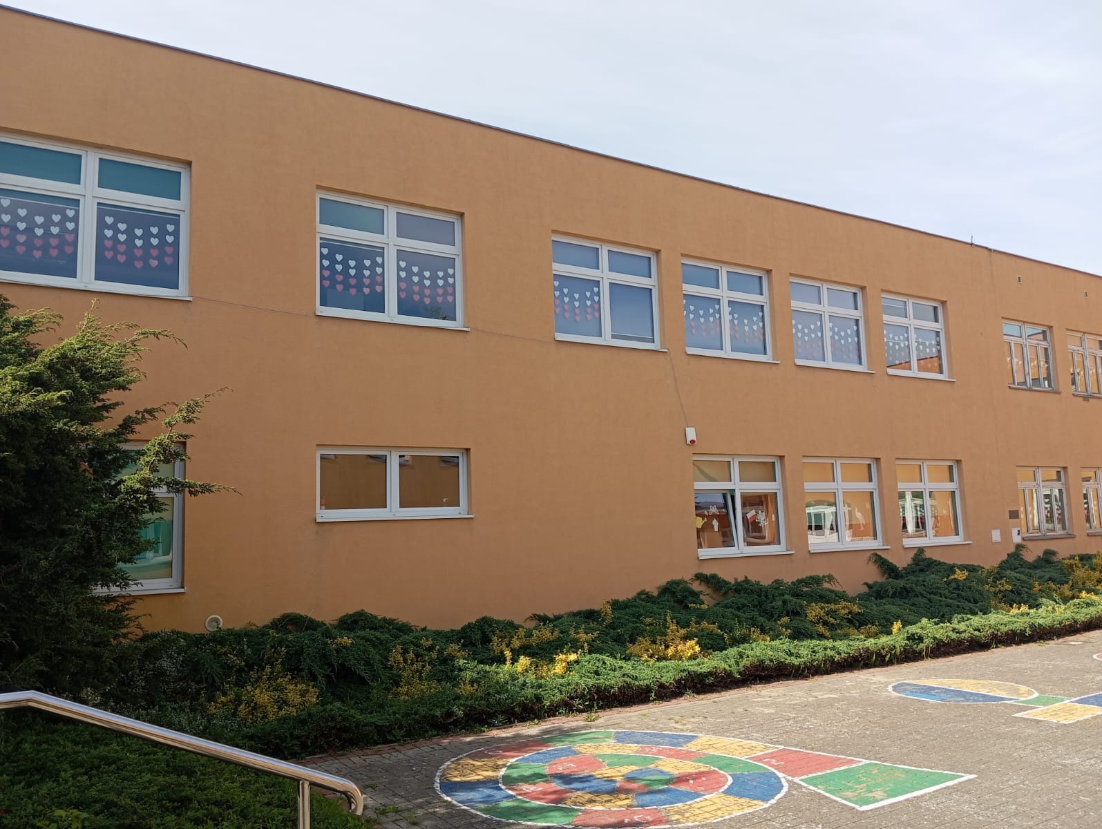 Fasada budynku w ciepłym, pomarańczowym odcieniu z licznymi białymi oknami, niektóre ozdobione papierowymi sercami, otoczona zielenią krzewów i drzew. Na pierwszym planie chodnik z namalowanymi...