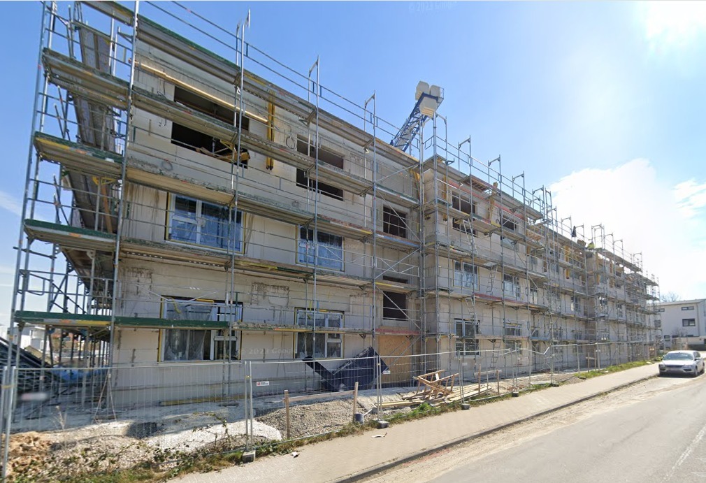 Wielopiętrowy budynek w trakcie budowy, z częściowo zamontowanymi oknami, otoczony rusztowaniami i ogrodzeniem tymczasowym, widoczny żuraw budowlany na dachu.