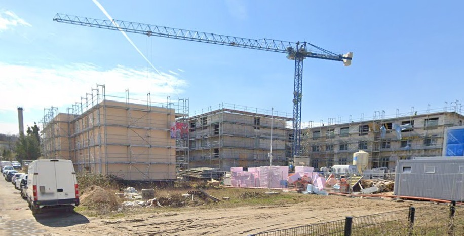 Budowa wielorodzinnego budynku z oknami w stanie surowym, rusztowania, żuraw budowlany i zaparkowane busy w słoneczny dzień.