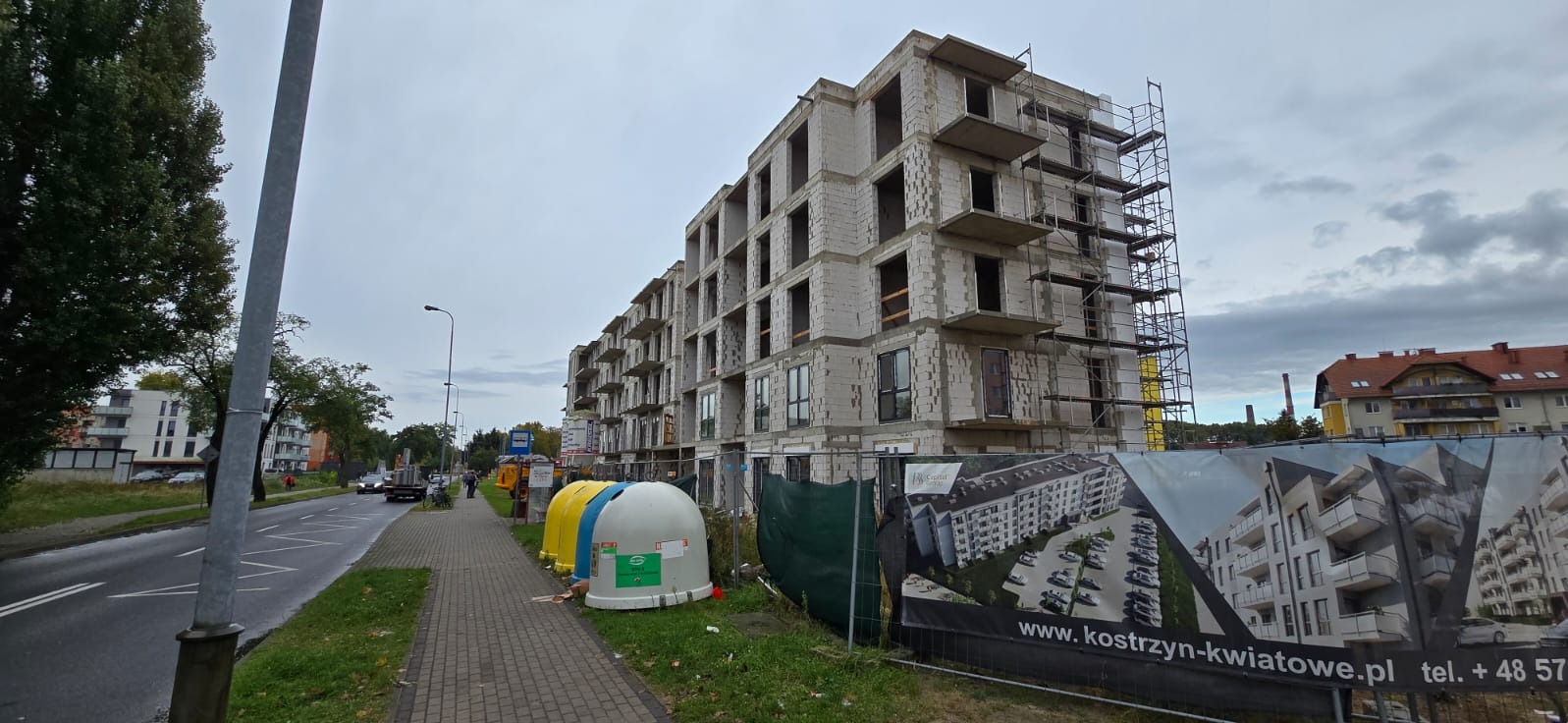 Surowy budynek wielorodzinny w trakcie budowy, widoczne rusztowania, otwory okienne i drzwiowe bez zamontowanej stolarki, obok ulicy stoją pojemniki na segregację odpadów, w tle inne budynki...