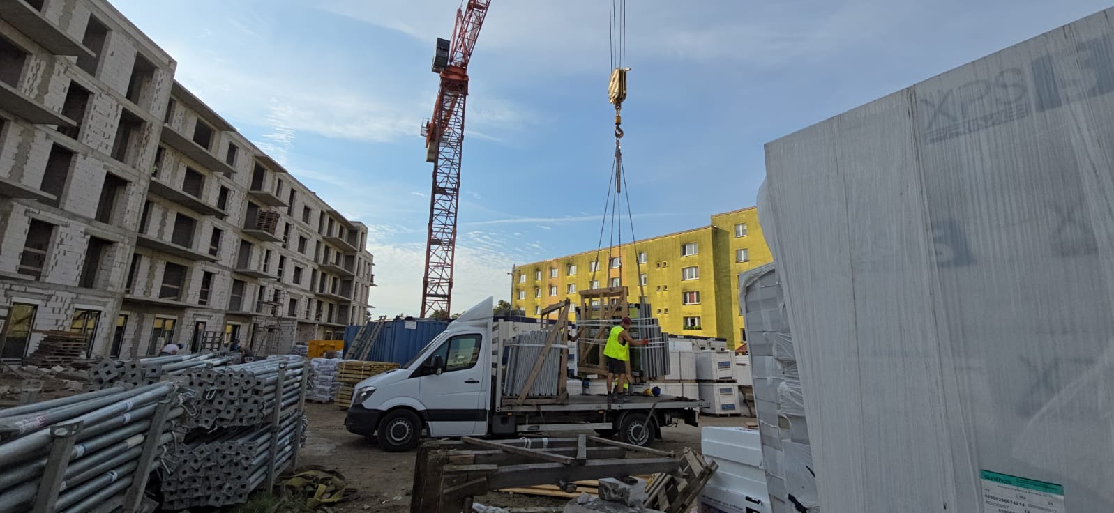 Rozładunek pakietu okiennych ram z ciężarówki za pomocą dźwigu na placu budowy, w tle budynek w stanie surowym i blok mieszkalny.