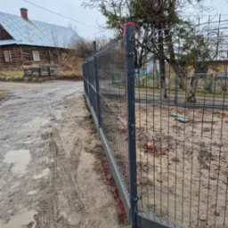 Fragment nowo postawionego ogrodzenia panelowego z podmurówką betonową, widoczne czerwone linki mocujące, błotniste podłoże i drewniany dom w tle.