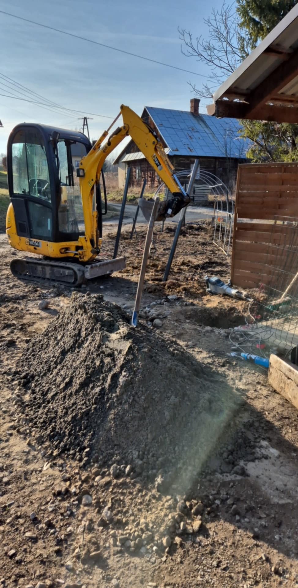 Żółta mini koparka przy pryzmie ziemi, w tle narzędzia i wykopany dół, widoczny fragment drewnianego płotu i budynek z niebieskim dachem.