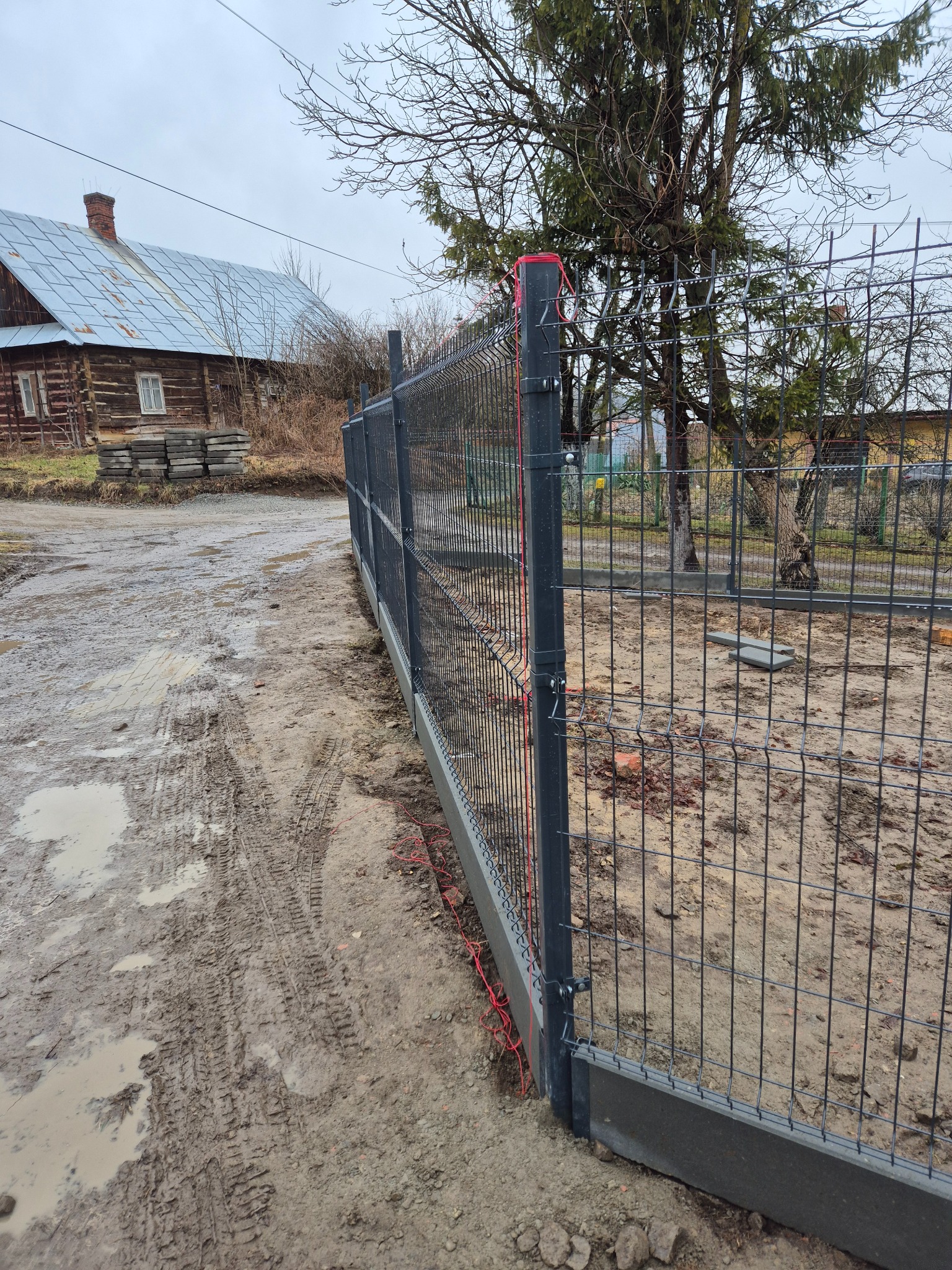 Fragment nowo postawionego ogrodzenia panelowego z podmurówką betonową, widoczne czerwone linki mocujące, błotniste podłoże i drewniany dom w tle.