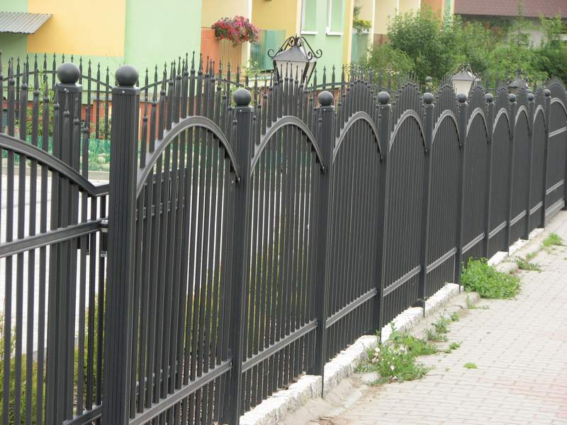 Czarne, metalowe ogrodzenie systemowe z dekoracyjnymi łukami i kulami na słupkach, oświetlone lampami w stylu retro, oddzielające posesję od chodnika wyłożonego kostką.