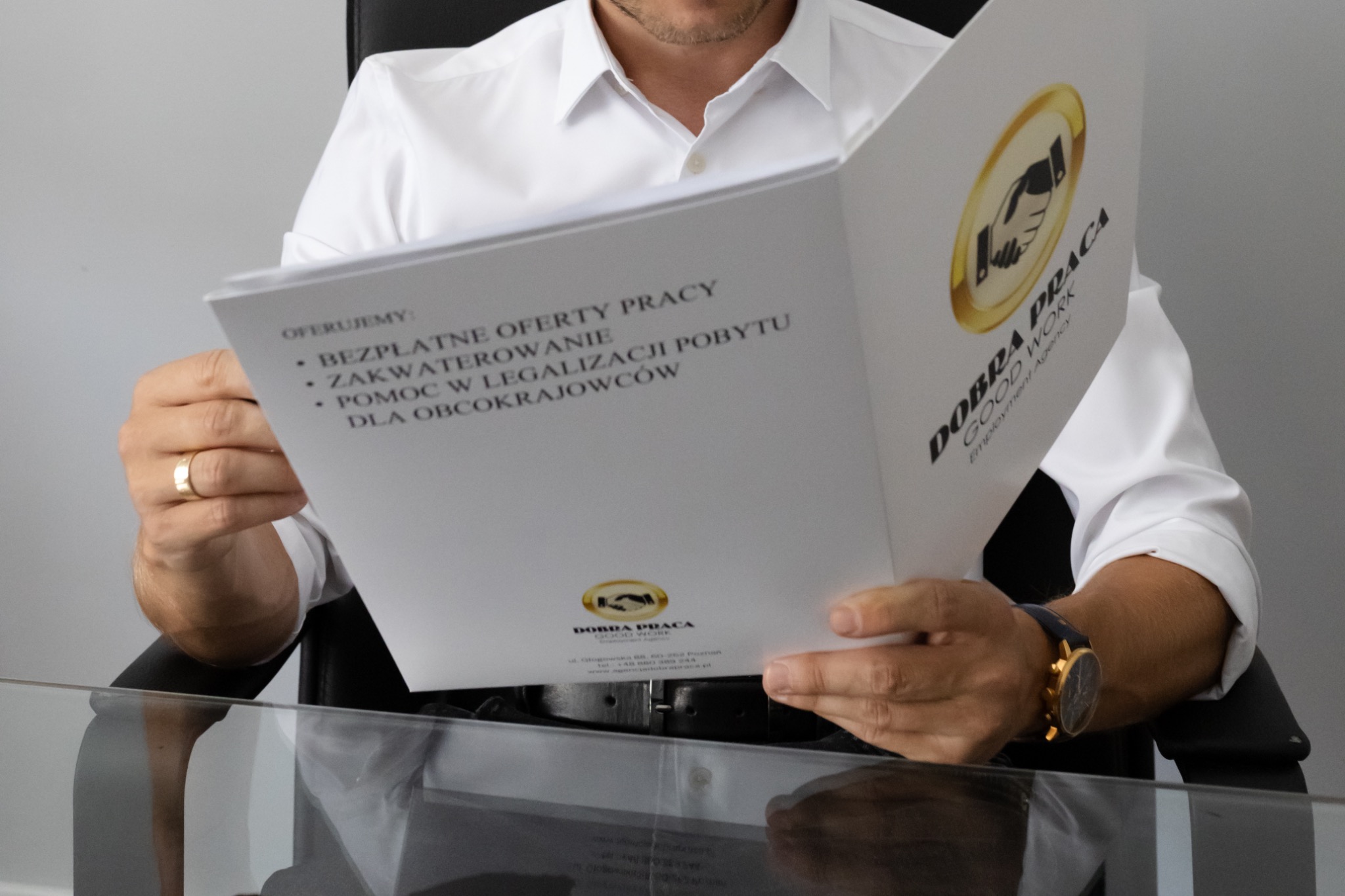 Mężczyzna w białej koszuli siedzi za biurkiem i przegląda otwarty folder z ofertą pracy dla obcokrajowców, prezentujący logo firmy rekrutacyjnej.