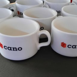 Koncept DTF - Nadruki na odzieży Nowy Sącz - Białe ceramiczne kubki z nadrukowanym logo firmy 'cano' z czerwonym symbolem i czarnym napisem, ustawione na szarym blacie.