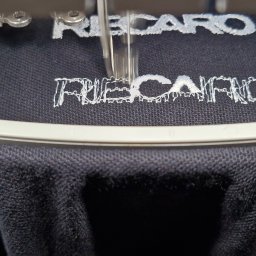 Koncept DTF - Nadruki na odzieży Nowy Sącz - Proces haftowania białego napisu 'RECARO' na czarnej koszulce za pomocą maszyny hafciarskiej, widoczne detale igły i tamborka.