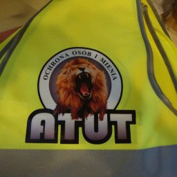 Koncept DTF - Nadruki na odzieży Nowy Sącz - Żółta kamizelka odblaskowa z nadrukowanym logo firmy ochroniarskiej: lew w okręgu z napisem 'OCHRONA OSÓB I MIENIA' oraz dużym napisem 'ATUT' poniżej.