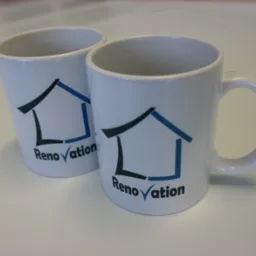 Dwa białe ceramiczne kubki z nadrukowanym logo firmy 'Renovation' w postaci uproszczonego domu z czarnym konturem i niebieskim dachem, ustawione na białym blacie.