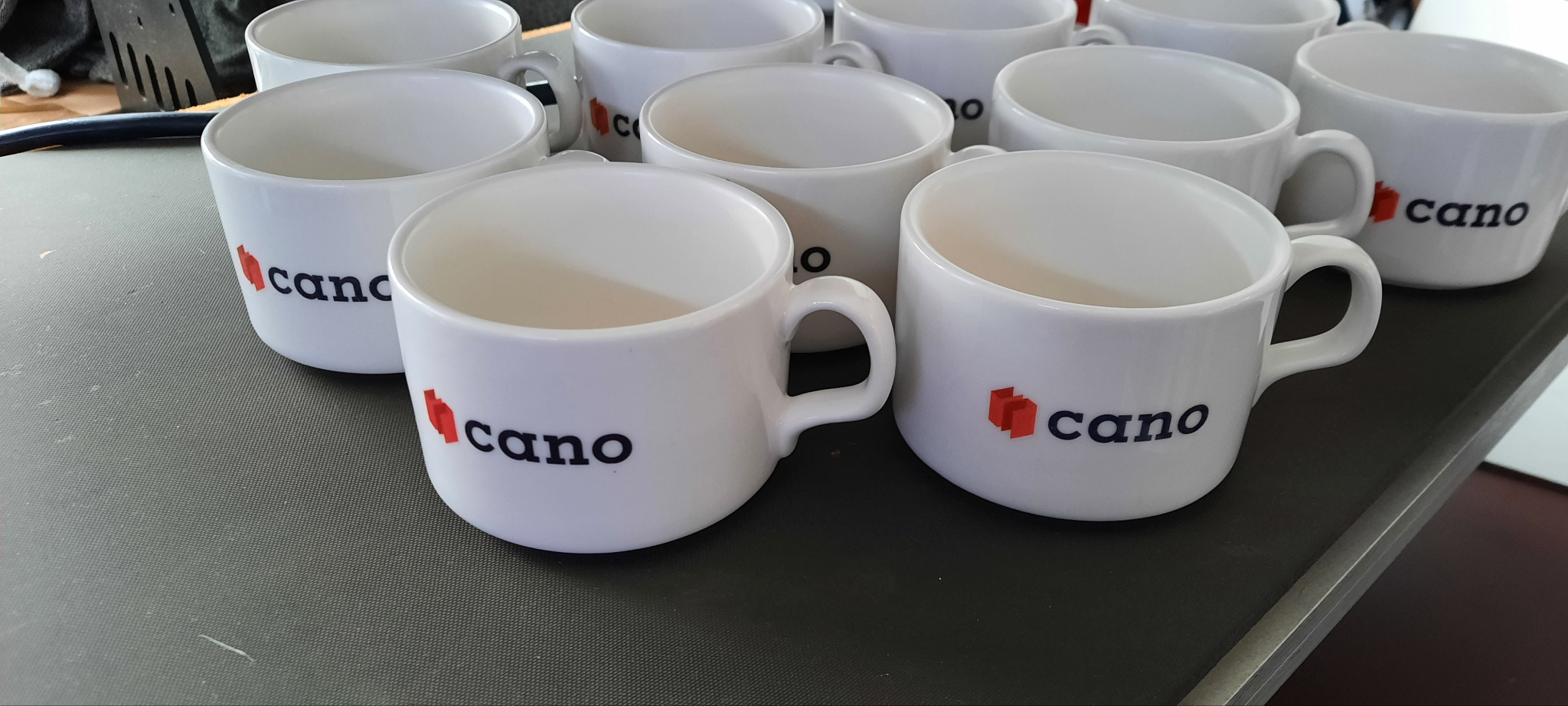 Białe ceramiczne kubki z nadrukowanym logo firmy 'cano' z czerwonym symbolem i czarnym napisem, ustawione na szarym blacie.