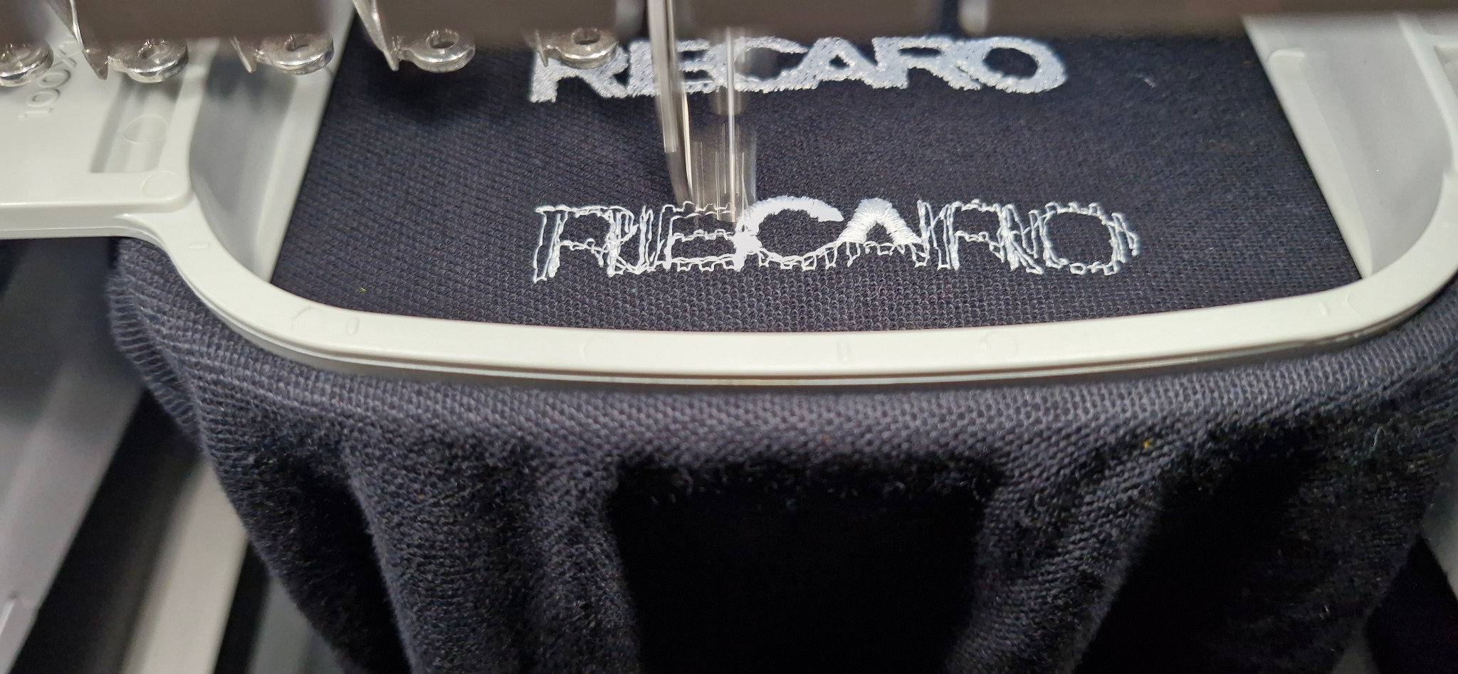 Proces haftowania białego napisu 'RECARO' na czarnej koszulce za pomocą maszyny hafciarskiej, widoczne detale igły i tamborka.