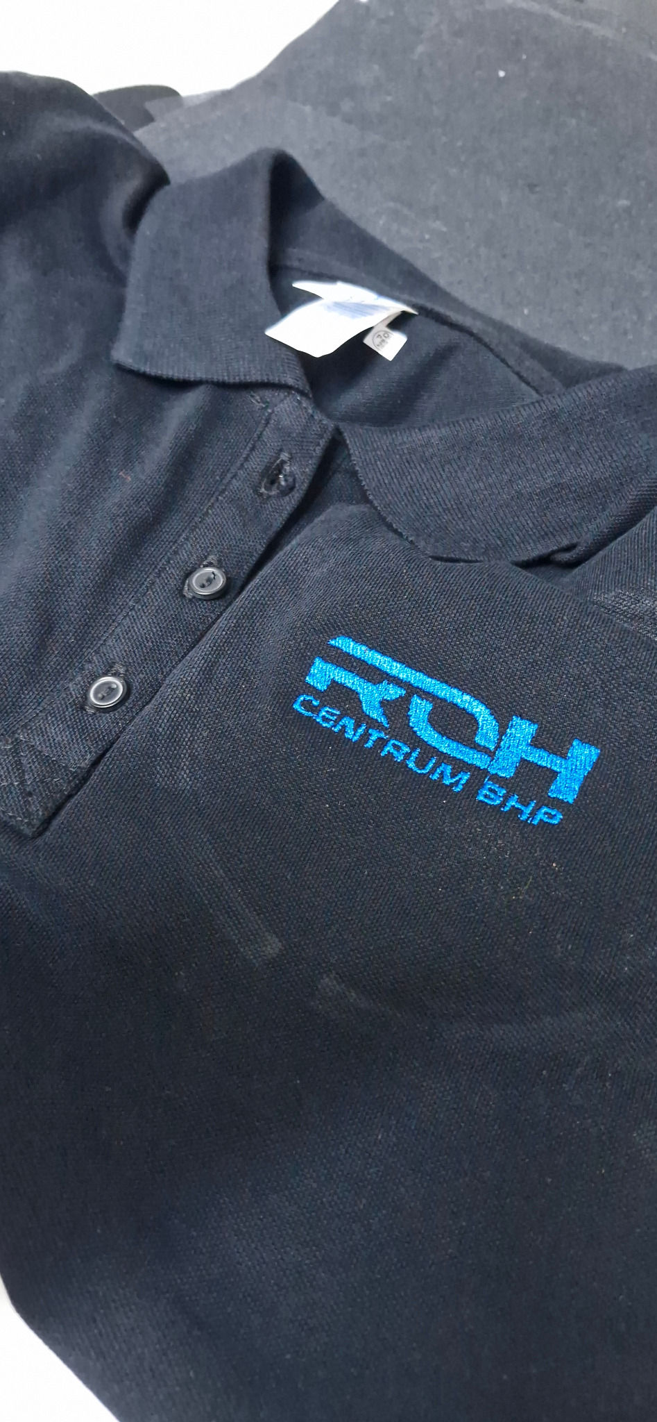 Ciemna koszulka polo z niebieskim haftowanym logo 'RDH CENTRUM BHP' na piersi, widoczne guziki i metka.