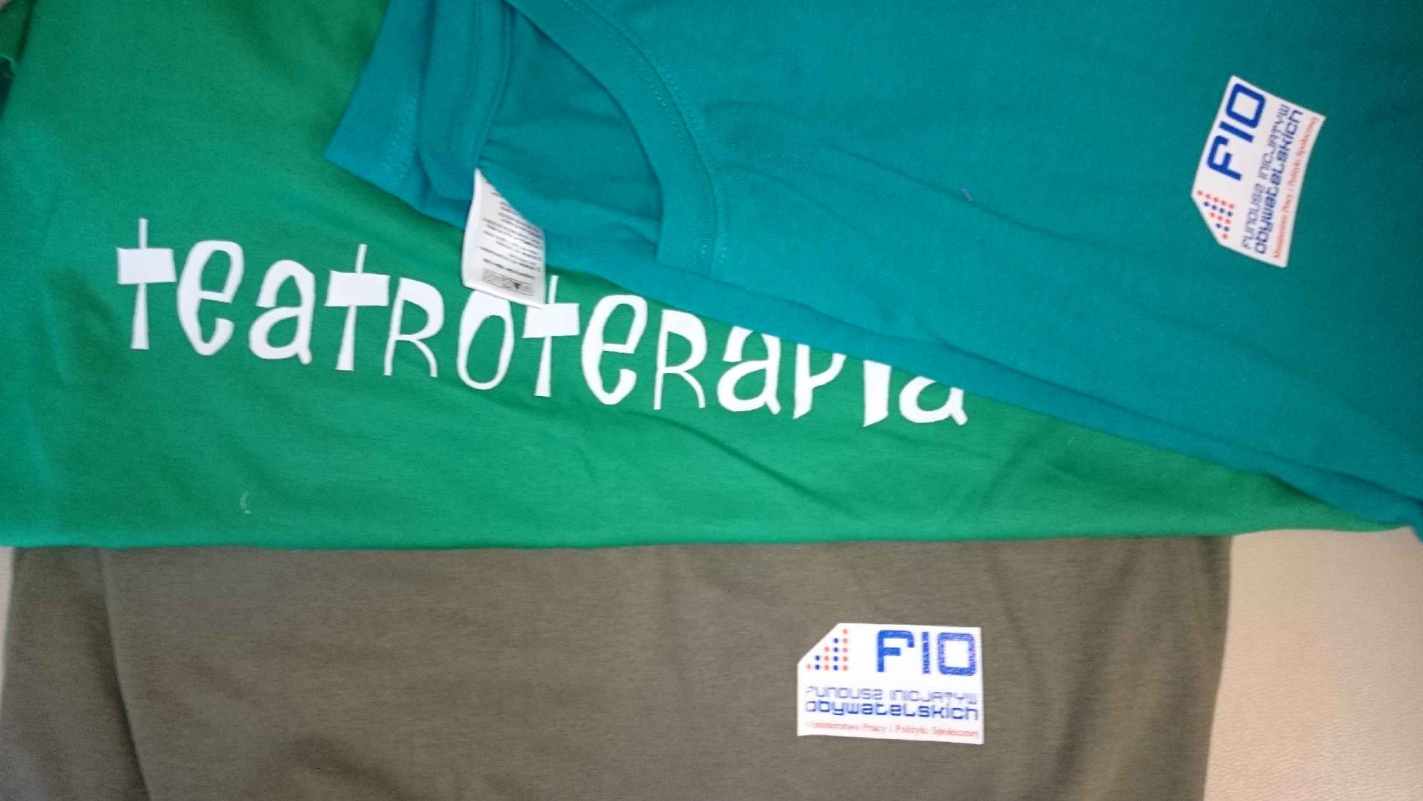 Stos trzech koszulek: zielonej z białym napisem 'teatroterapia', turkusowej i szarej, obie z logo FIO Funduszu Inicjatyw Obywatelskich.