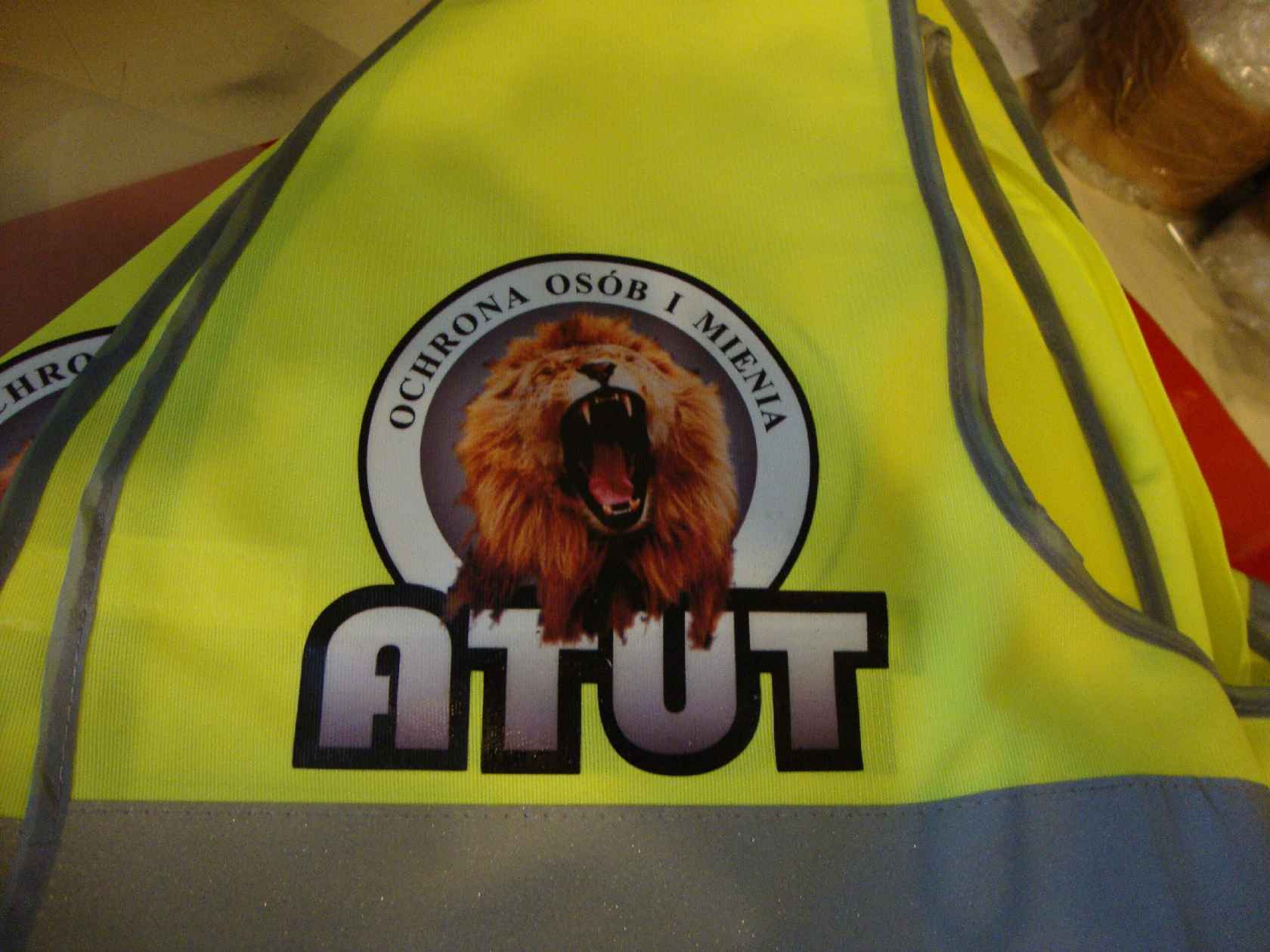 Żółta kamizelka odblaskowa z nadrukowanym logo firmy ochroniarskiej: lew w okręgu z napisem 'OCHRONA OSÓB I MIENIA' oraz dużym napisem 'ATUT' poniżej.