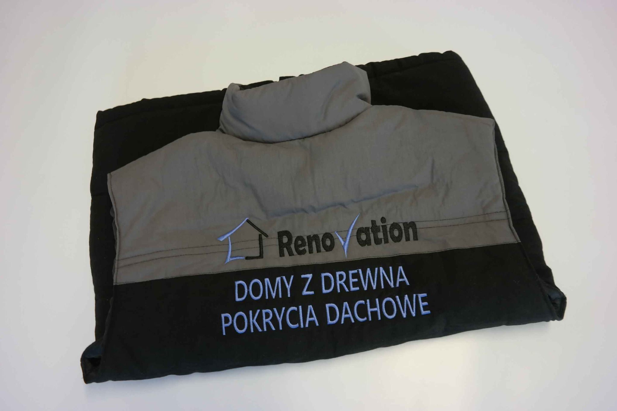 Czarno-szara torba robocza z wyhaftowanym logo firmy budowlanej 'Renovation' oraz napisem 'Domy z drewna, pokrycia dachowe' w kolorze niebieskim, leżąca na białym tle.