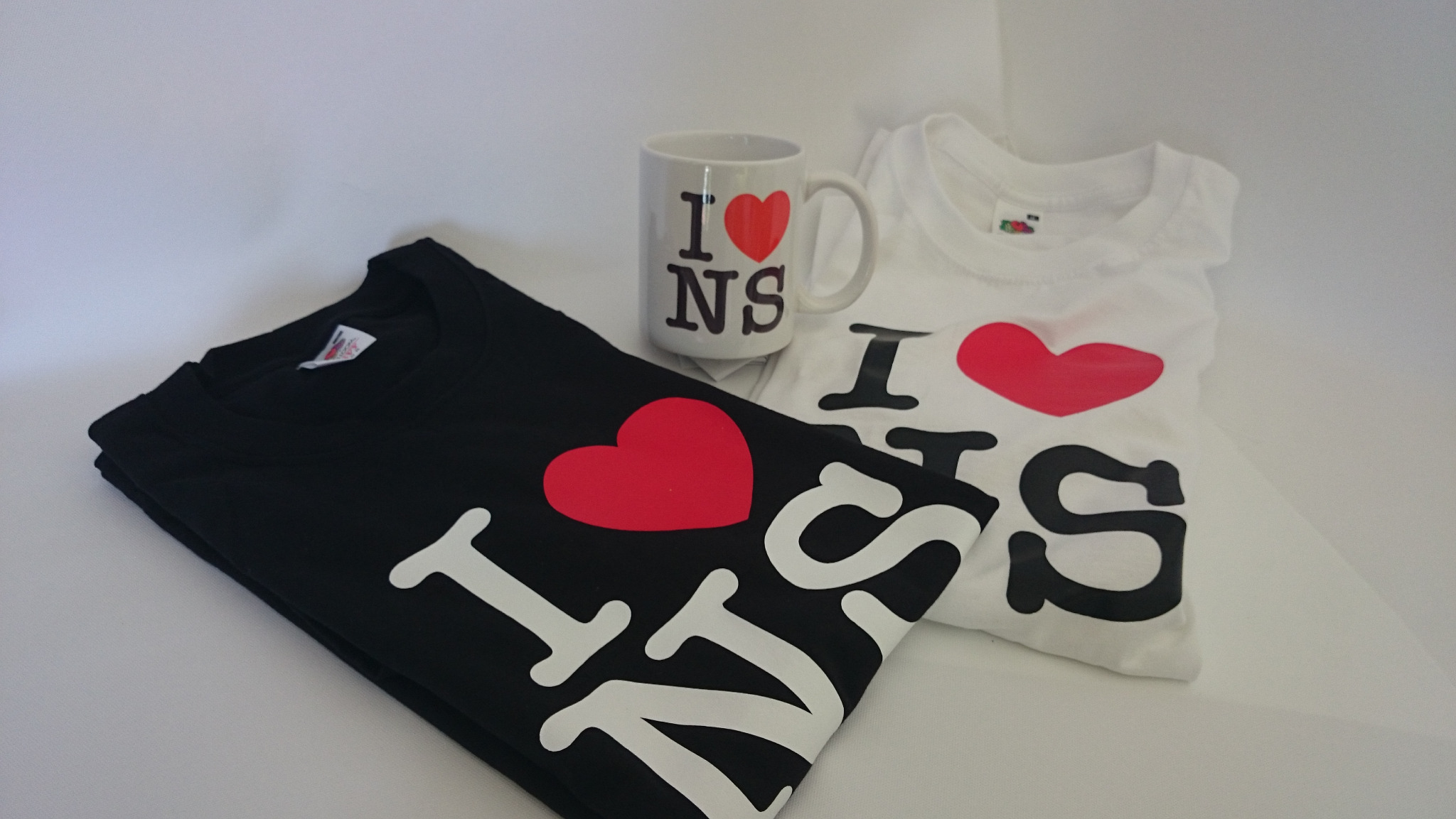 Dwie koszulki, czarna i biała, z motywem 'I ❤️ NS' oraz kubek z tym samym nadrukiem, ułożone na białym tle. Koszulka czarna jest złożona, a biała częściowo rozłożona.