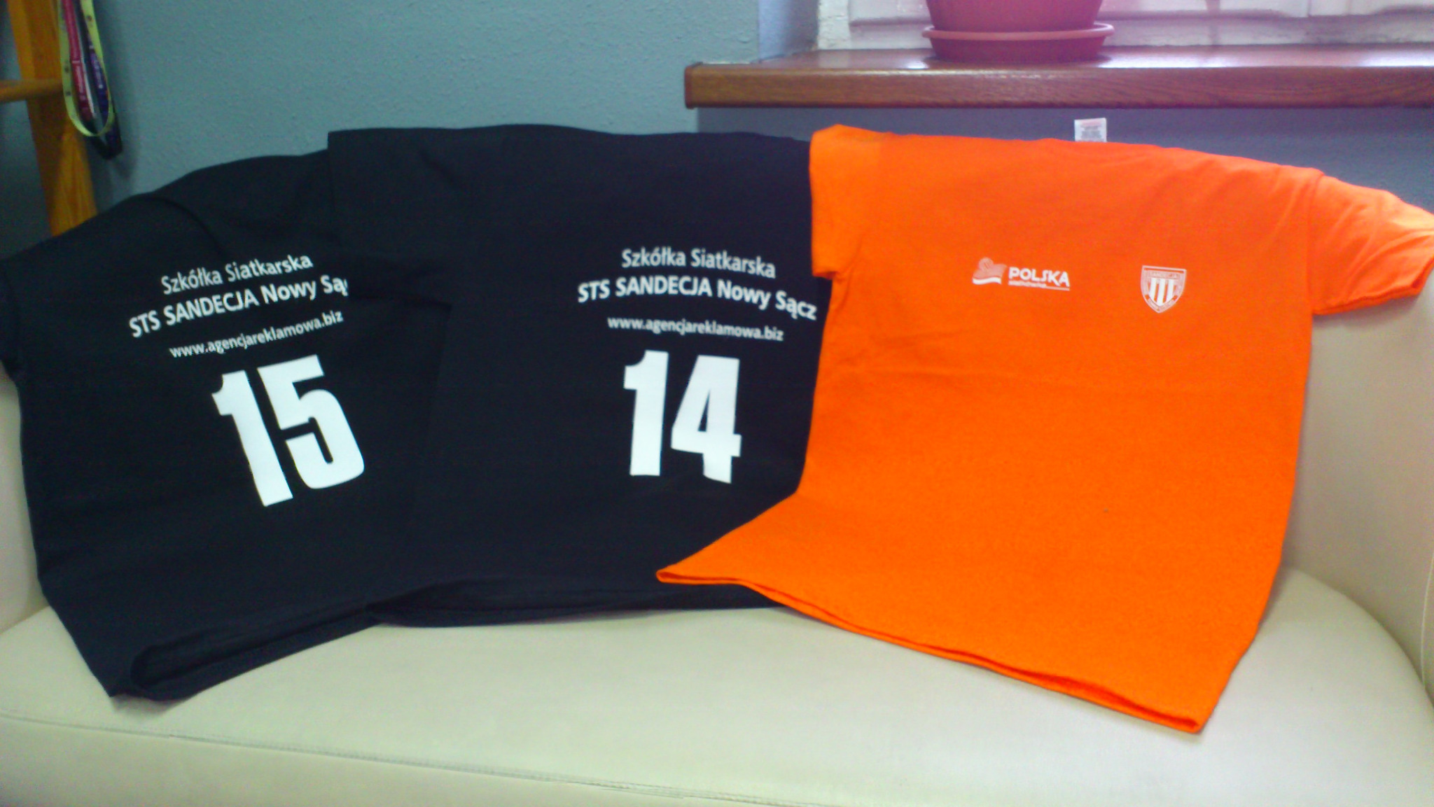 Dwa czarne T-shirty z białym nadrukiem 'Szkółka Siatkarska STS SANDECJA Nowy Sącz' i numerami 15 i 14, obok pomarańczowy T-shirt z logo i napisem 'POLSKA', leżące na białej kanapie.