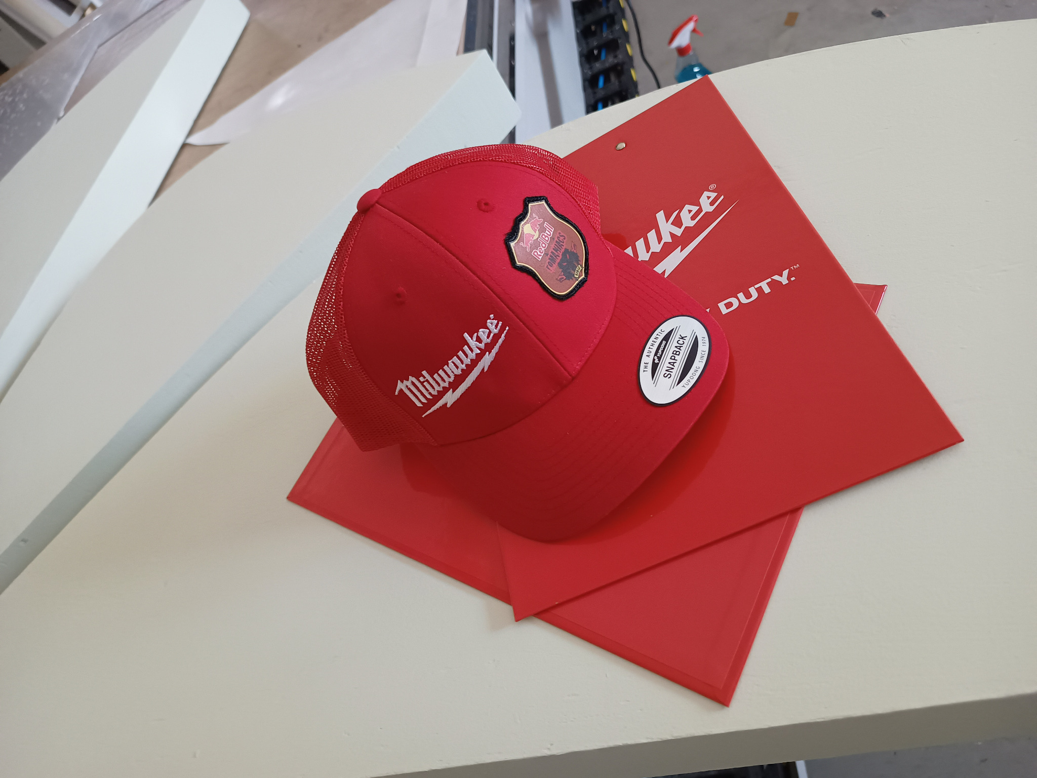 Czerwona czapka z daszkiem Milwaukee z naszywką Red Bull Tomakawks leżąca na czerwonej teczce z logo Milwaukee, na jasnym stole.
