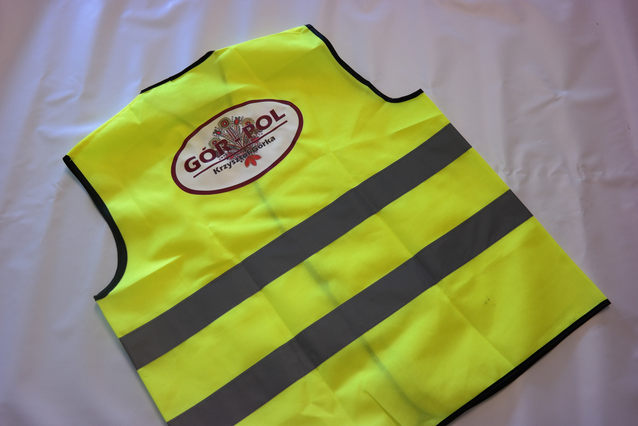 Żółta kamizelka odblaskowa z trzema szarymi pasami i logo 'GÓR-POL Krzysztof Górka' z motywem kwiatowym, leżąca na białym, pogniecionym tle.