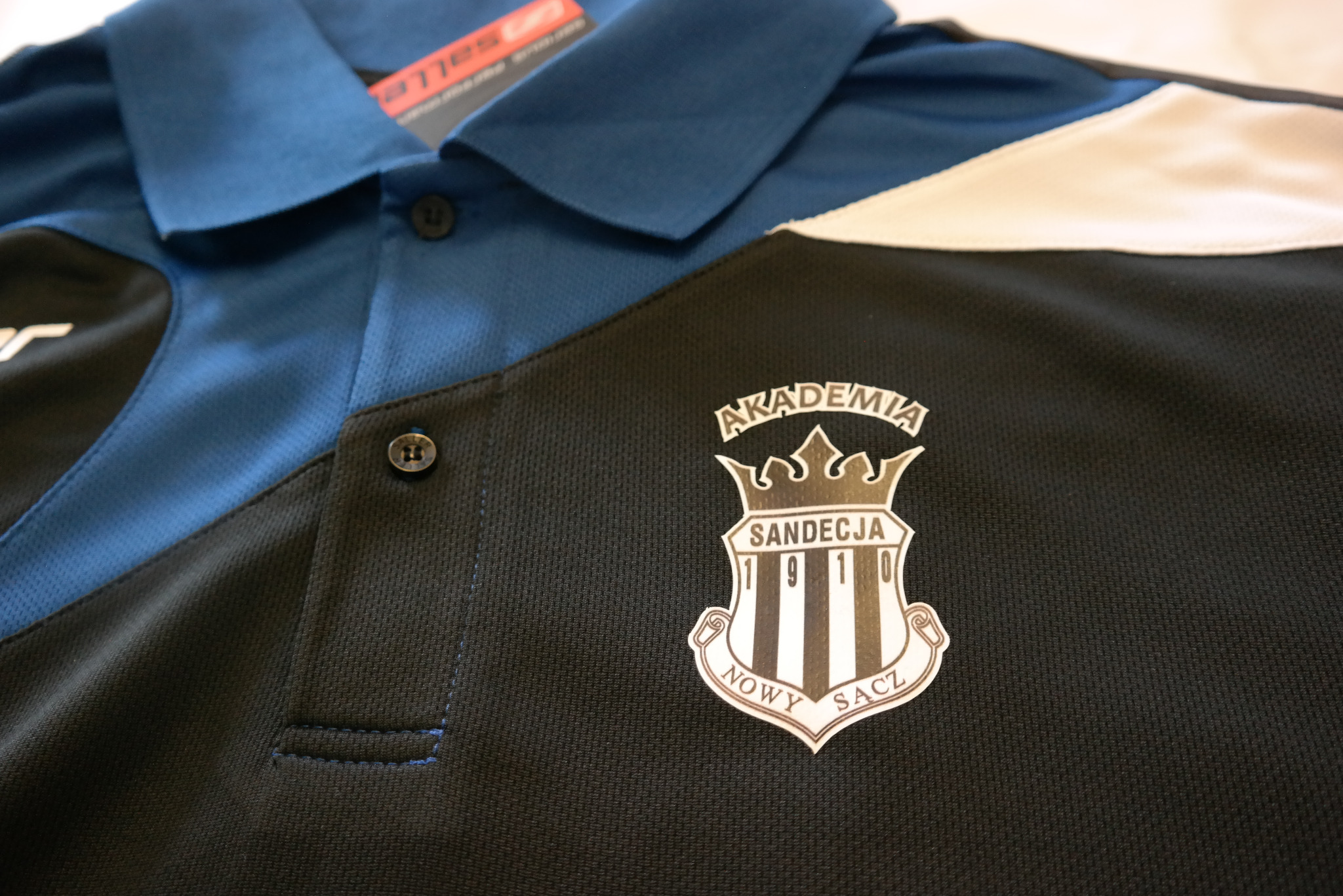 Koszulka polo w kolorach czarnym, niebieskim i białym, z herbem 'Akademia Sandecja Nowy Sącz 1910' po prawej stronie.