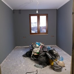 Perfect Home - Pokój w trakcie malowania na szaro, zabezpieczona podłoga folią, widoczny stos worków z materiałami, wiadro z farbą i butelki z wodą.