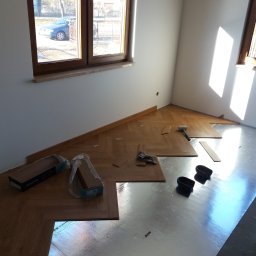 Perfect Home - Układanie parkietu w jodełkę w jasnym pomieszczeniu z oknami, widoczne paczki paneli Berry Alloc, podkład pod panele, klej i gumowy młotek.