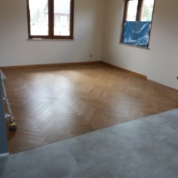 Perfect Home - Jasne, przestronne wnętrze z nową podłogą ułożoną w jodełkę, kontrastujące szare płytki podłogowe w przejściu, okno zabezpieczone folią malarską, drabina malarska w rogu.