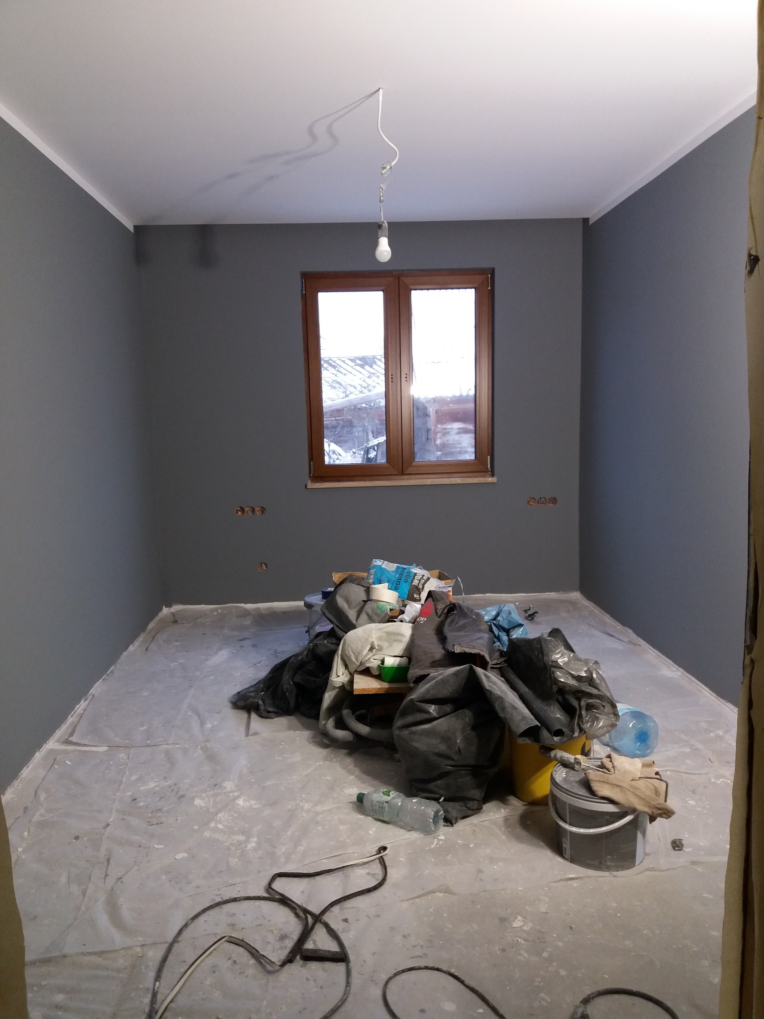 Pokój w trakcie malowania na szaro, zabezpieczona podłoga folią, widoczny stos worków z materiałami, wiadro z farbą i butelki z wodą.