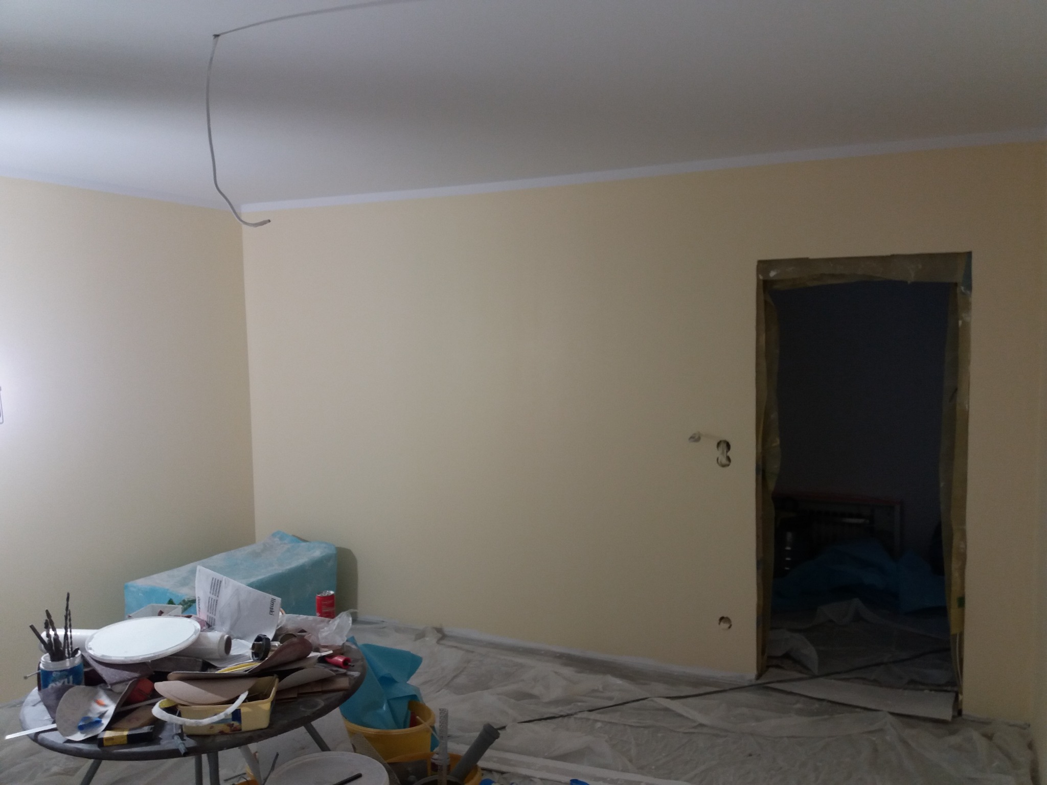 Pokój w trakcie malowania na kremowo, widoczne narzędzia malarskie na stoliku, zabezpieczona folią podłoga i futryna drzwi, wystający kabel z sufitu.