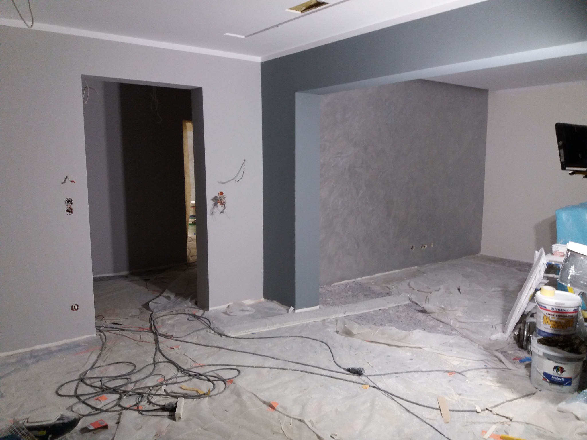 Wnętrze w trakcie malowania, ściany w odcieniach szarości, zabezpieczona folią podłoga, widoczne puszki z farbą i narzędzia malarskie.