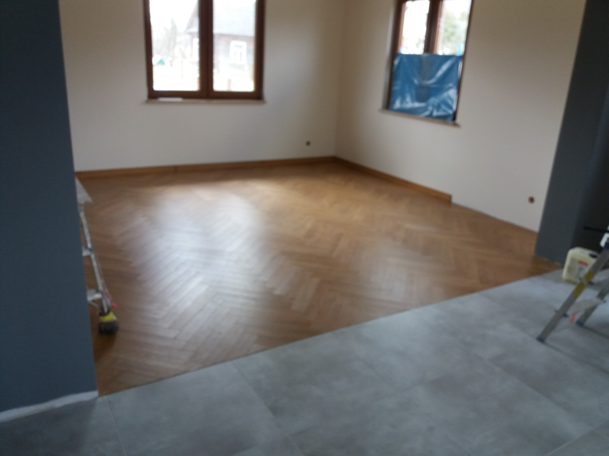 Jasne, przestronne wnętrze z nową podłogą ułożoną w jodełkę, kontrastujące szare płytki podłogowe w przejściu, okno zabezpieczone folią malarską, drabina malarska w rogu.