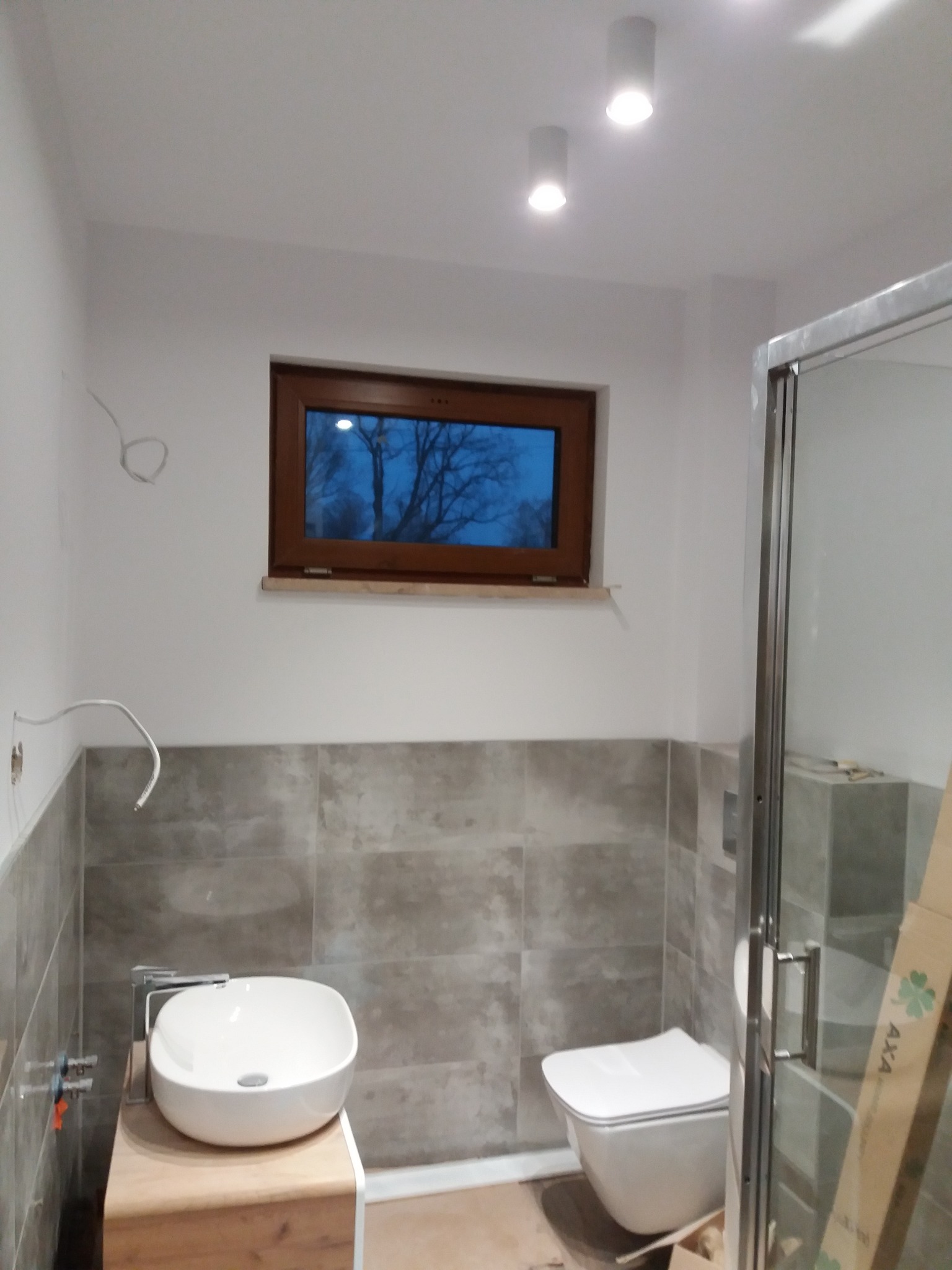 Nowoczesna łazienka po renowacji z umywalką nablatową na drewnianej szafce, wiszącą toaletą i kabiną prysznicową z widocznym kartonem producenta.