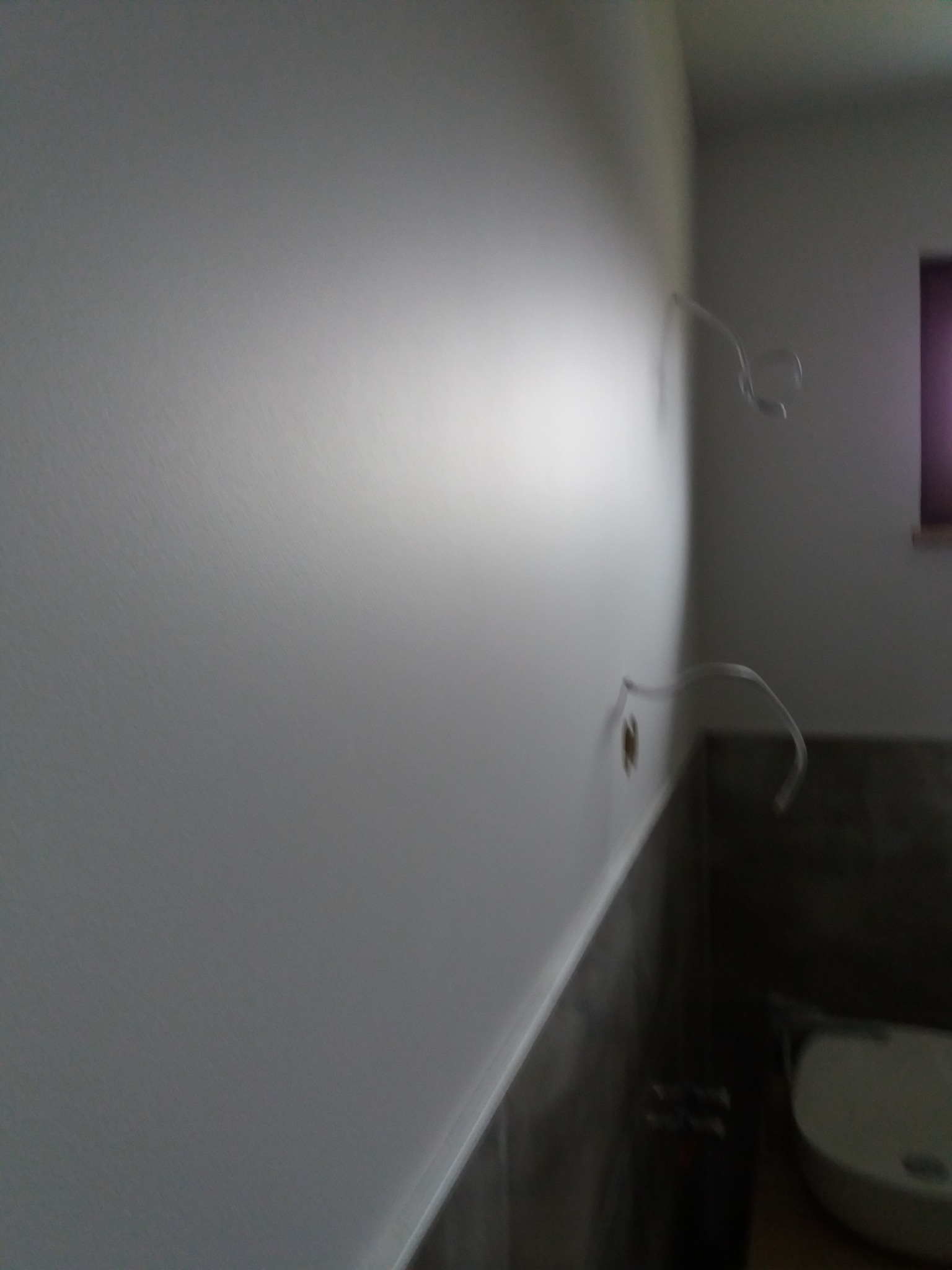 Świeżo pomalowana, gładka ściana w łazience z wystającymi przewodami elektrycznymi przed montażem osprzętu, widoczne szare płytki u dołu ściany.