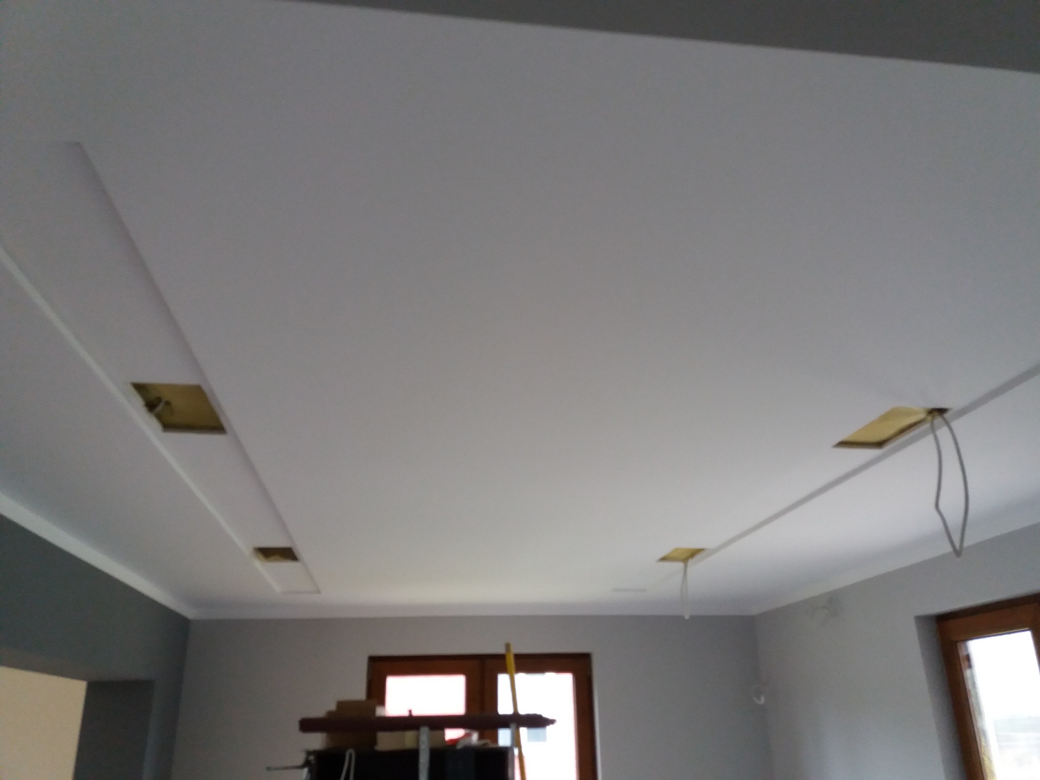 Podwieszany sufit z otworami na oświetlenie, widoczne kable, ściany pomalowane na szaro, częściowo widoczne okna i drzwi.