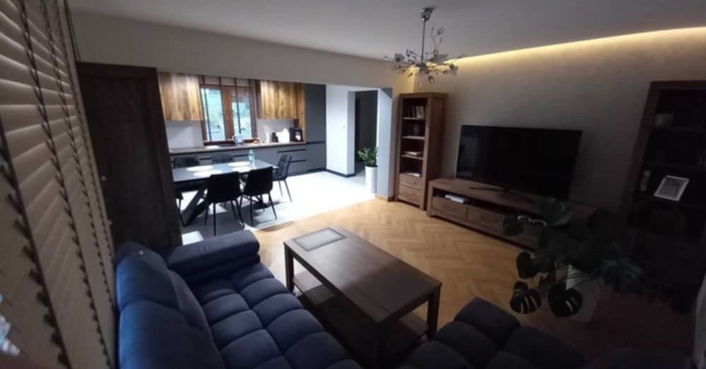 Kompleksowo wykończone wnętrze: salon z aneksem kuchennym, drewniana podłoga w jodełkę, niebieska sofa, drewniane meble i oświetlenie LED podsufitowe.