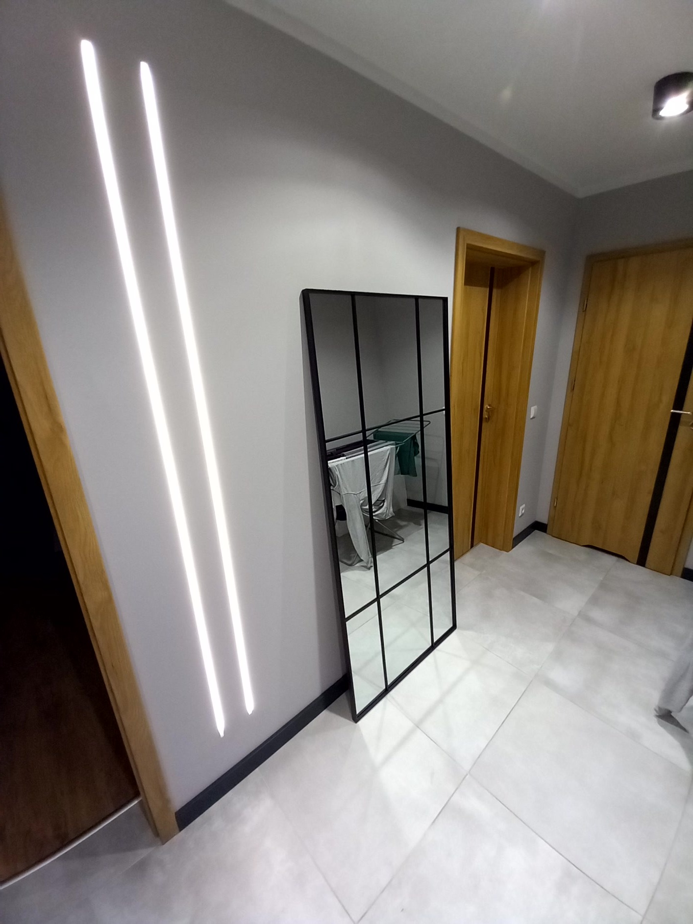 Nowoczesne wnętrze z lustrem w czarnej ramie, odbijającym suszarkę na pranie, oświetlone pionowymi lampami LED na szarej ścianie i jasną podłogą z płytek.