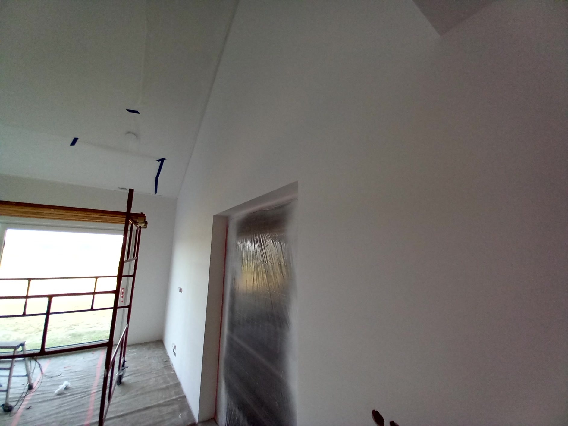 Wnętrze w trakcie malowania na biało, zabezpieczone folią drzwi, widoczne rusztowanie przy oknie i taśmy malarskie na suficie.