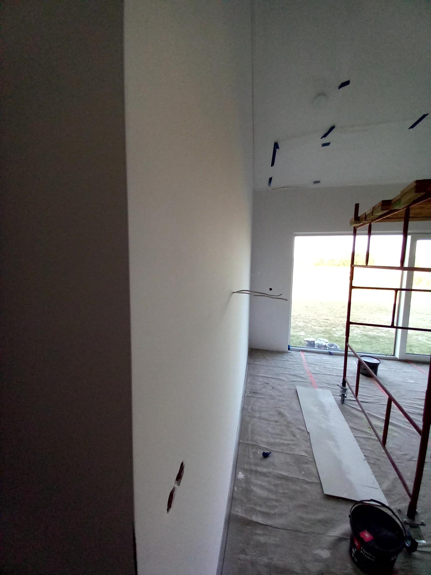 Wnętrze w trakcie prac malarskich: świeżo pomalowana, jasna ściana z widocznymi przewodami elektrycznymi, zabezpieczona podłoga z rozłożoną folią ochronną, rusztowanie malarskie i wiadro z farbą...