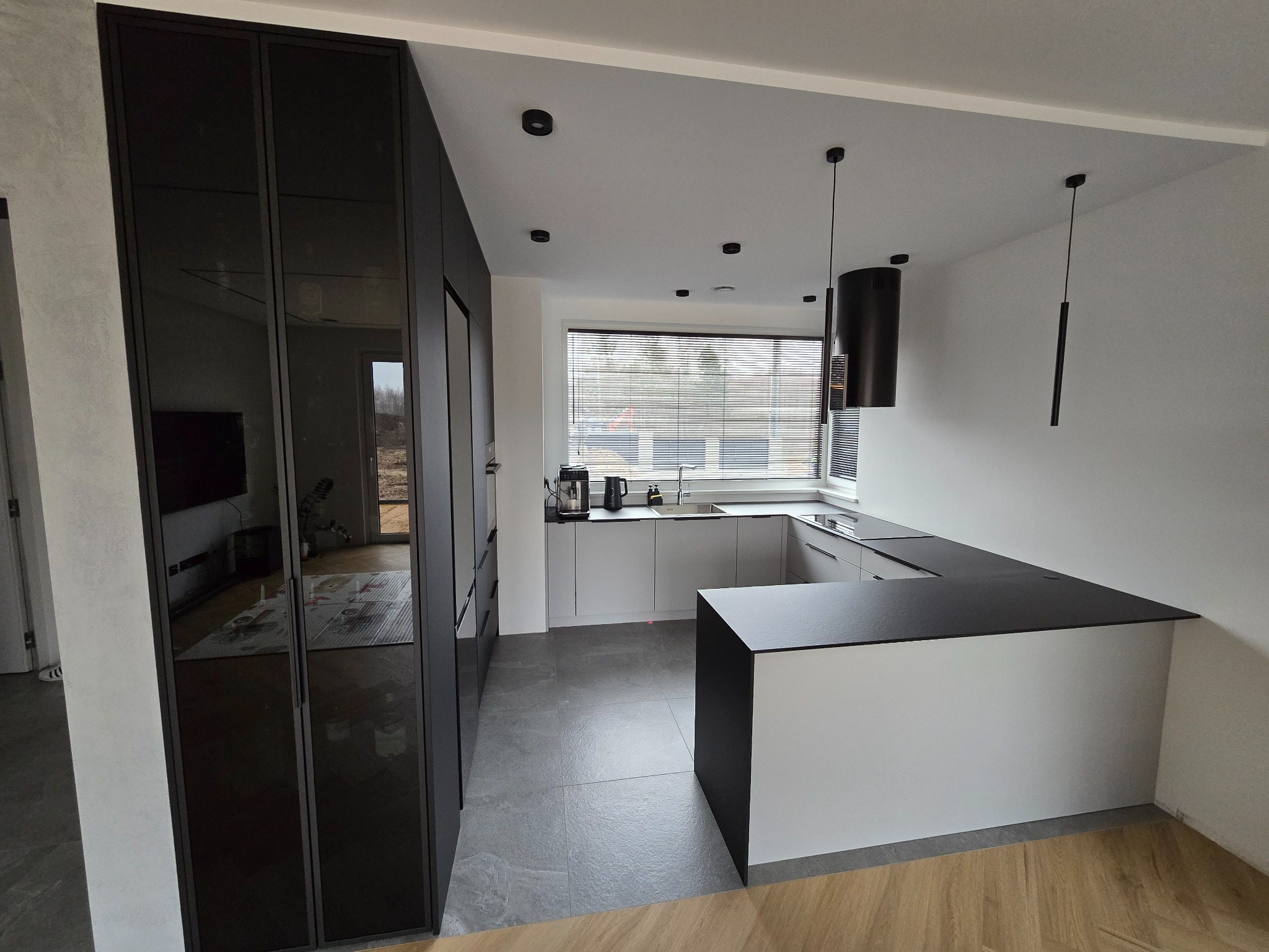 Nowoczesna kuchnia z szarą zabudową, czarnym blatem i szafą wnękową z czarnymi, lustrzanymi drzwiami, oświetlona wiszącymi lampami.