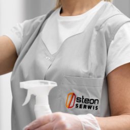 STEON OCHRONA Sp. z o.o. - Pracownica w szarym uniformie z logo firmy 'Osteon Serwis' trzyma biały spryskiwacz, dłoń w przezroczystej rękawiczce, przygotowana do pracy.