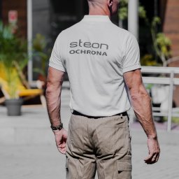 STEON OCHRONA Sp. z o.o. - Mężczyzna tyłem w koszulce z napisem 'STEON OCHRONA' i spodniach typu cargo, stojący na zewnątrz budynku.