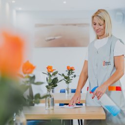 STEON OCHRONA Sp. z o.o. - Pracownica w szarym uniformie z logo firmy spryskuje płynem czyszczącym blat stołu z bukietami pomarańczowych róż w wazonach, w tle jasne wnętrze biura z obrazem.